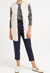 Vitt fuskpälsväst utan ärmar över en grå stickad tröja, matchat med marinblå cropped-byxor och vita slip-on skor.