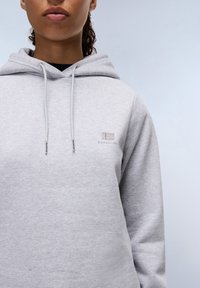 Sudadera gris claro con capucha ajustable. Presenta un pequeño logo bordado en el pecho. Fabricada con una tela suave y texturizada.