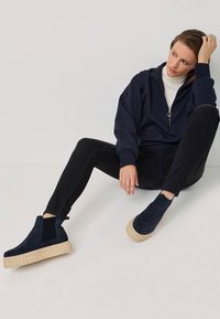 Bocanci navy din suede, cu panouri laterale stretch și tălpi din cauciuc chunky bej, asortați cu blugi negri și un hanorac navy cu fermoar.