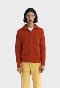 Rood gebreid jack met ritssluiting, hoge kraag en geribbelde textuur. Heeft diagonaal gestikte naden en een aansluitende tailleband, gecombineerd met gele broek.