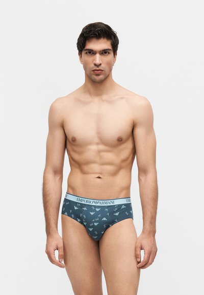 Άνδρας χωρίς πουκάμισο όρθιος, φοράει μπλε εσώρουχα Emporio Armani με λογότυπο, ουδέτερη έκφραση, απλό ανοιχτό φόντο.