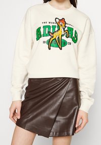 Krämfärgad sweatshirt med grön och svart branding, försedd med en broderad rendesign; kombinerad med en brun fusk läder omslagskjol.