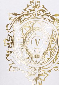 Biały materiał z dużym złotym metalicznym logo, zdobionym misternymi wzorami kwiatowymi, napisem "Versace Jeans Couture" oraz wyraźnym centralnym "V".