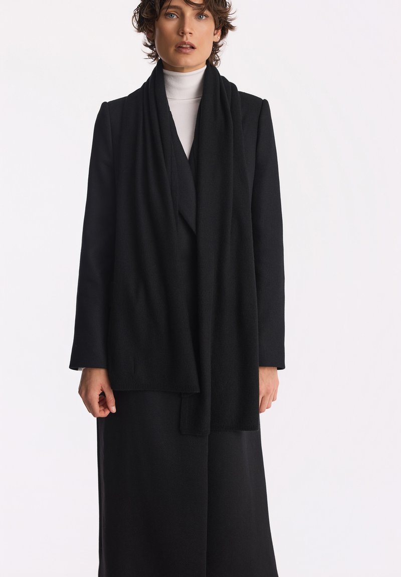 Czarny płaszcz z wełny z domieszką, z drapowanym przodem, długimi rękawami i otwartym krojem. Nosi się go na białym golfie, o minimalistycznej strukturze.