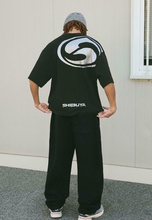 Crna oversized majica s srebrnim kružnim logotipom na leđima i tekstom "SHIBUYA" na dnu. Uparena s tamnoplave trenirke i tenisicama.