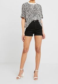 Personne portant un t-shirt blanc à imprimé léopard noir, un short noir taille haute et des sandales blanches à talons hauts, debout sur un sol gris.