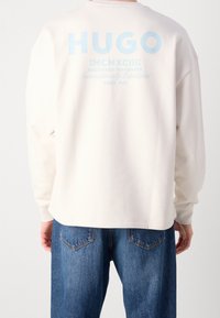 Persona che indossa una felpa crema con la scritta blu "HUGO MCMXCIII Registered Trademark Since 1993" sulla schiena e jeans di denim blu.