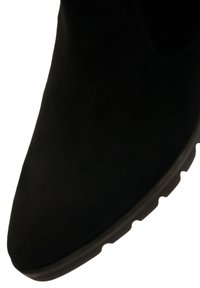 Close-up of a black suede shoe toe with a textured rubber sole edge visible on a white background.