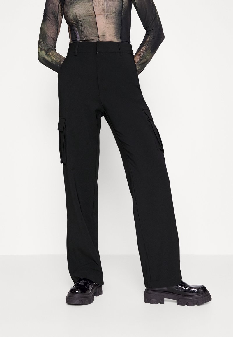 Personne portant un pantalon cargo noir taille haute avec poches latérales et des chaussures noires à plateforme épaisse, debout les mains dans le dos.