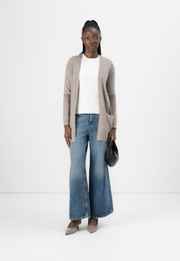 Kvinna som står klädd i beige karderoll, vit topp, vida jeans, beige klackar och håller en liten svart väska mot en vit bakgrund.