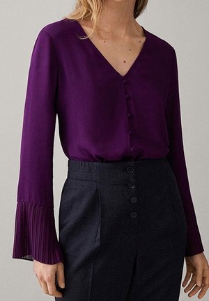 Kobieta ubrana w fioletową bluzkę z długim rękawem z plisowanymi mankietami i dekoltem w serek, zestawioną z wysokim stanem szarych spodni z guzikami.