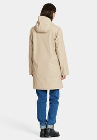 Beige vedenpitävä takki hupulla, suora leikku ja pitkät hihat. Yhdistetty sinisiin farkkuihin ja mustiin nilkkureihin.