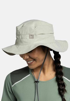 Smiling person with braided hair wearing light beige wide-brim sun hat with chin strap and green athletic shirt with dark shoulder panels.