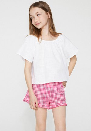 Jeune fille debout, les mains sur les hanches, portant un chemisier blanc à manches courtes et un short rose à carreaux avec des ourlets à volants.