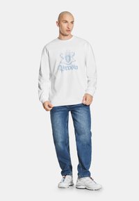 Biała bawełniana koszulka z długim rękawem z niebieskim wzorem graficznym na klatce piersiowej, w połączeniu z niebieskimi dżinsami i białymi butami sportowymi.