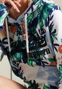 Persona que lleva una sudadera blanca con patrones florales en verde y naranja y el texto negro "Superdry" en el pecho, atada con cordones blancos.