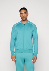 Teal zip-up športna jakna z belimi akcenti in logotipom na prsih. Ima rebrast ovratnik in manšete, v kompletu s usklajenimi hlačami.