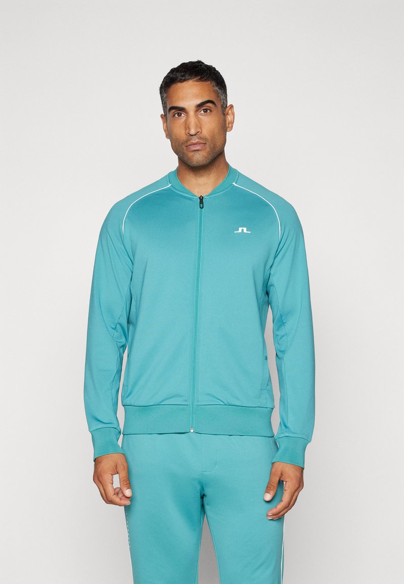 Teal zip-up športna jakna z belimi akcenti in logotipom na prsih. Ima rebrast ovratnik in manšete, v kompletu s usklajenimi hlačami.