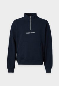 Marineblå fleece-pullover med halv lynlås og høj krave, der har en front kangaroo-lomme og "GUESS JEANS"-logo i hvid broderi.