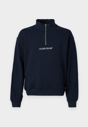 Marineblå fleece-pullover med halv lynlås og høj krave, der har en front kangaroo-lomme og "GUESS JEANS"-logo i hvid broderi.