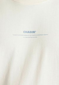 T-shirt color crema con maniche corte e scollo a costine. Presenta il testo azzurro chiaro "CHASIN'" e una frase descrittiva sotto. Tessuto morbido.