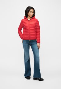 Γυναίκα που φοράει κόκκινο πουπουλένιο μπουφάν με κουκούλα, μπλε τζιν παντελόνι bootcut και μαύρα παπούτσια, στέκεται με το ένα χέρι στην τσέπη της σε λευκό φόντο.