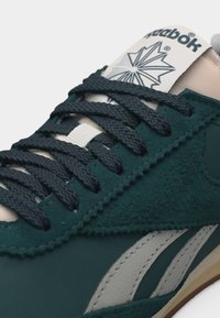 Σκούρο πράσινο αθλητικό παπούτσι Reebok με μαύρα κορδόνια, μπεζ γλώσσα και λευκό λογότυπο Reebok στο πλάι και στη γλώσσα.