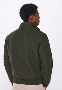 Olive green bomber jacket with a high collar, ribbed cuffs, and hem. Features a smooth texture and minimal seam detailing on the back.