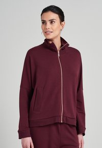 Burgundská zipová bunda z měkkého materiálu, s vysokým límcem, postranními kapsami a uvolněným střihem s dlouhými rukávy.