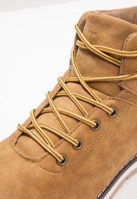 Bota de ante marrón con cordones trenzados amarillos y rojos, ojales de metal y puntera redonda. Presenta un collar acolchado y una suela de goma blanca.