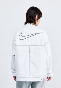 Fehér Nike dzseki, melyen egy nagy fekete Swoosh logó és a 95-ös szám található a hátán. Az anyag sima textúrájú, kontrasztos varrással.