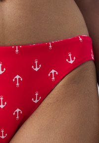 Rdeča bikini spodnja dela z majhnim belim vzorcem sidra, gladka tekstura tkanine in nizkim dizajnom na bokih.