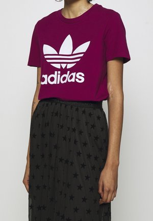 Personne portant un t-shirt violet Adidas avec un logo blanc et une jupe noire à motif d’étoiles, debout devant un fond uni.