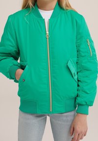 Henkilö, jolla on päällään kirkkaan vihreä vetoketjullinen bomber-takki, jossa on ribbaus hihansuissa ja vyötäröllä, vaaleat farkut sekä valkoinen paita alla.