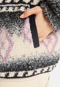 Pullover in pile caratterizzato da una superficie decorata con colori grigio, crema e rosa. Include una tasca laterale con una finitura nera testurizzata.