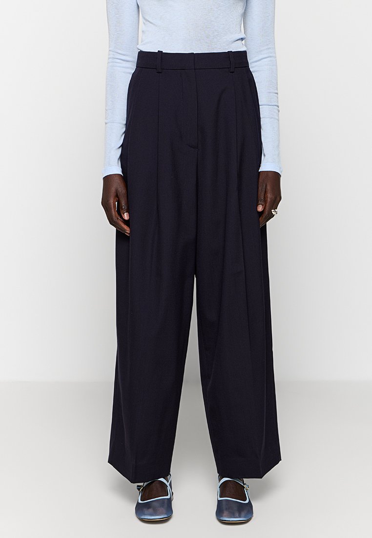 Granatowe spodnie z szerokimi nogawkami, fałdami, wysokim stanem i paskiem. Gładka tkanina o wyraźnej strukturze, zestawione z jasnoniebieskimi butami.