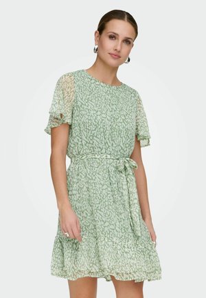 Kvinne iført lys grønn kjole med kort erme, mønstret, med rysjekant nederst og knyttet midje, tilbehør med sølv ringøredobber.