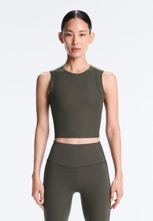 Kvinde med håret sat op i en knold iført en ærmeløs, olivengrøn ribbet crop top og matchende højtaljede leggings mod en ensfarvet baggrund.