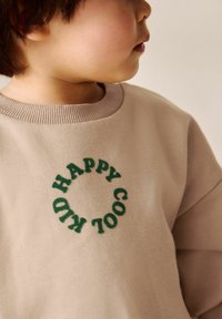 Ein kleines Kind trägt einen beigen Sweatshirt mit grüner, kreisförmiger Aufschrift "HAPPY COOL KID" und schaut seitlich vor einem schlichten Hintergrund.