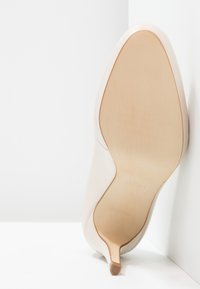 Scarpa beige con tacco alto posizionata con la suola rivolta verso l'alto contro un angolo bianco, mostrando la suola liscia e la punta del tacco sottile.