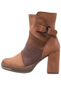 Botte de cheville en suède marron avec un talon bloc, un design superposé dans des nuances de marron, et une boucle décorative avec un accent métallique circulaire.