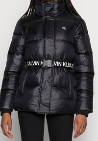 Černá prošívaná bunda s vysokým límcem, polstrovaným designem a logem Calvin Klein. Má kontrastní pásek a boční kapsy. Hladká, lesklá textura.