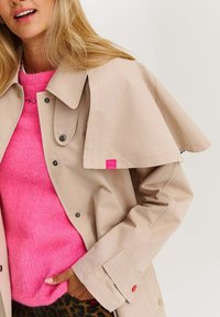 Žena v béžovém trenčkotě na jasně růžovém svetru, s rukou v kapse leopardích kalhot, usmívající se proti jednoduchému pozadí.