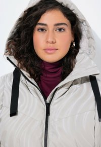 Chaqueta acolchada en gris claro con un patrón de ondas brillante, que cuenta con capucha, cremalleras negras y correas ajustables en el cuello.