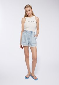 Top senza maniche bianco con la scritta "Venice Beach", abbinato a pantaloni corti di jeans azzurri a vita alta. Calzature: ciabatte blu.