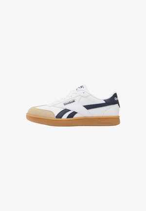 Zapatilla blanca Reebok con puntera beige, rayas azul marino y suela de goma, vista de lado.