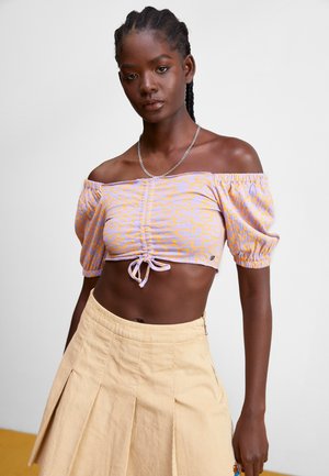 Top court à épaules dénudées en lilas avec un motif floral jaune ; il présente des manches bouffantes et un nœud à l'avant. Associé à une jupe en corduroy beige.