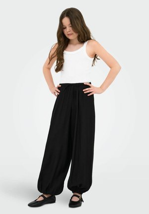 Jente med langt brunt hår iført en hvit ermeløs topp, løse svarte bukser med elastiske mansjetter og svarte sko, står med hendene på hoftene.