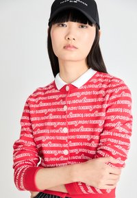 Crvena cardigan s ponavljajućim bijelim uzorkom "Tommy Jeans" loga, ovratnikom s bijelim obrubom i kopčanjem na gumbe sprijeda.