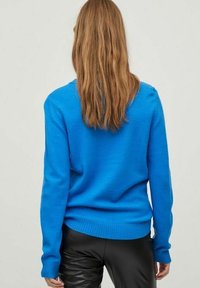 Mujer con cabello largo castaño claro, vestida con un suéter azul brillante y pantalones de cuero negros, vista de espaldas sobre un fondo liso.